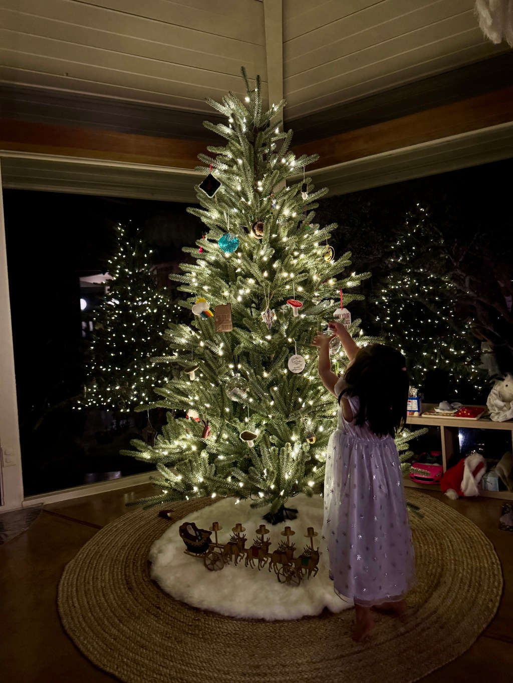 A young girl in a cozy, minimalist home carefully placing ornaments on a glowing Christmas tree. The warm light of the tree softly illuminates the room, creating a serene and festive atmosphere.